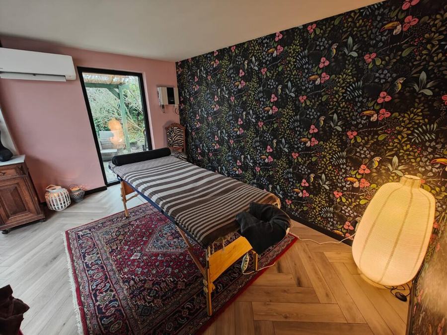De behandelkamer: een massagetafel met gestreept doek op een Perzisch tapijt, donker bloemenbehang en een papieren lamp